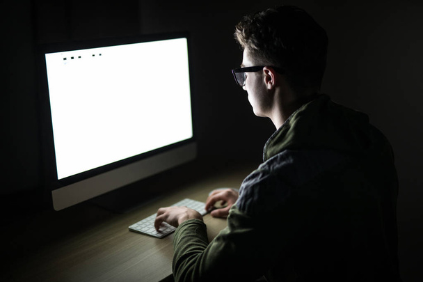 Крупним планом молодий чоловік сидить і використовує порожній екранний комп'ютер у темній кімнаті. хакер або програміст у темній кімнаті
. - Фото, зображення