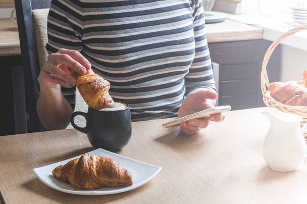Breakfast: The girl dips a croissant in a mug with coffee, and in the other hand holds a smartphone. Morning bright sun. - Photo, image