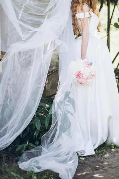 stijlvolle bruid met pioenroos boeket en lange sluier, poseren in zonnige tuin op trouwdag. luxe bruiloft meisje, gelukkig jonggehuwde in zijden jurk. romantische momenten - Foto, afbeelding