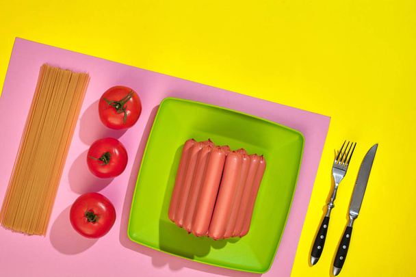 Много сырых сосисок на тарелке. На желтом фоне с макаронами и овощами, вид сверху
. - Фото, изображение