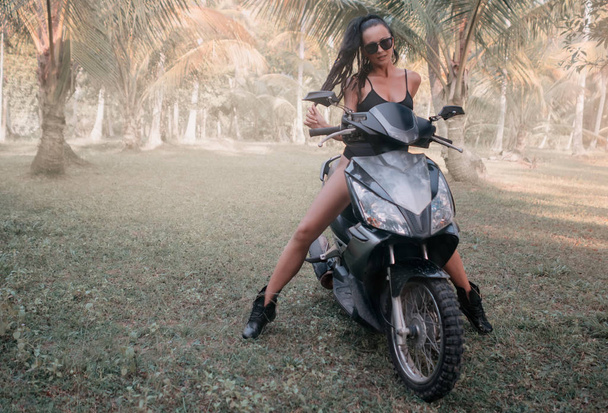 Beautiful, sexy young brunette in sunglasses is sitting on a scooter in jungle - Фото, зображення