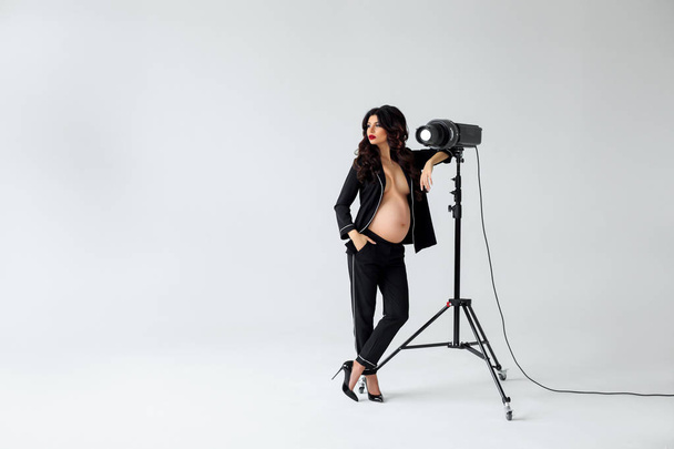 Ready for perfect shot. Beautiful young pregnant woman in black silk pajama looking away while leaning to lightning tool against white background - Photo, Image