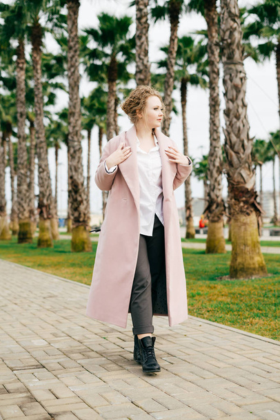 stylish curly young woman in a pink coat walking in the park, against the backdrop of palm trees, warm spring weather - Photo, Image