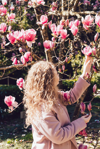 curly young woman walking in the park on a sunny day, standing next to a pink magnolia - Zdjęcie, obraz