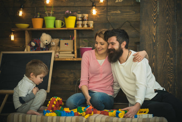 Happy parents hug each other while looking at son. Family playing with plastic blocks on sofa. Side view concentrated boy plays with toy train - Fotografie, Obrázek