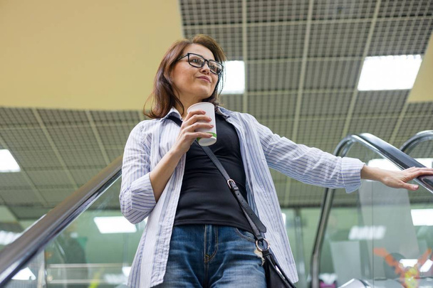 Middle-aged woman with cup of coffee, background shopping mall entertainment center. Lifestyle, leisure and people concept. - Foto, afbeelding