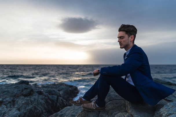 Tired young man in elegant suit sits on the background of the sea and sky - Fotoğraf, Görsel