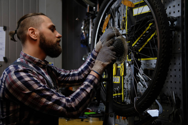 Красивый стильный мужчина в фланелевой рубашке и джинсовом комбинезоне, работающий с велосипедным колесом в мастерской
. - Фото, изображение
