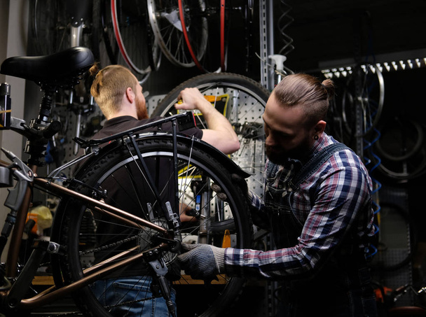 zwei gut aussehende Männer, die mit einem Fahrrad in einer Werkstatt arbeiten. Arbeiter reparieren und montieren Fahrrad in Werkstatt. - Foto, Bild