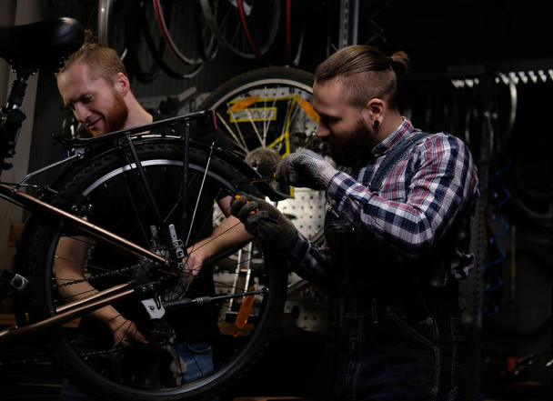 Dwóch przystojnych mężczyzn stylowe, praca z rowerem w warsztacie. Pracowników naprawy i uchwyty rowerowe w warsztacie. - Zdjęcie, obraz