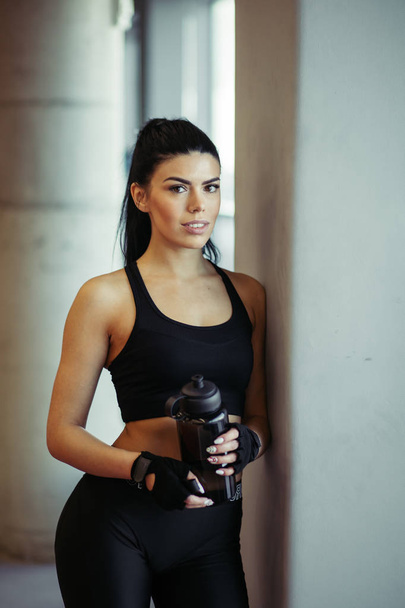 Female fit model posing with water after training near windows in gym - Фото, зображення