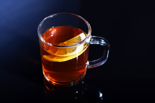 A glass mug of tea with lemon stands on a black glass. A cup of tea is beautifully reflected in the glass - Foto, afbeelding