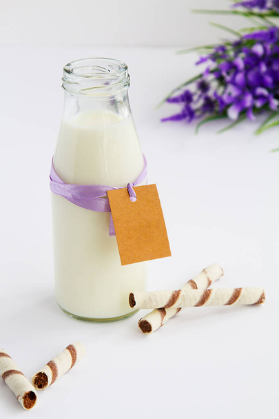 botella de leche con etiqueta para escribir deseos, junto a sus tubos de oblea de chocolate sobre fondo blanco y flores de color lila lado derecho
 - Foto, imagen