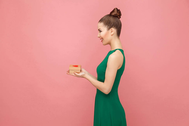 side view of smiling beautiful woman in green dress presenting gift box with heart on pink background - 写真・画像