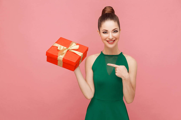woman holding red gift box and pointing finger at it while posing on pink background - Photo, image