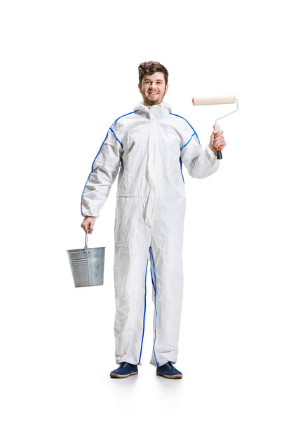 Young male decorator painting with a paint roller climbed a ladder isolated on white background. - 写真・画像