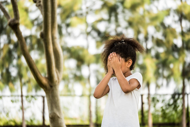 Uma menina está de pé em um parque, cobrindo seu rosto com as mãos
 - Foto, Imagem