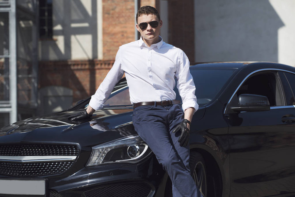  Young handsome man is standing at an black car on the background of a city building - Photo, Image