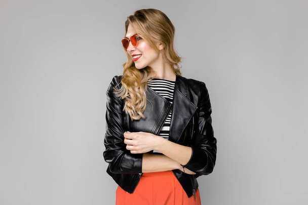 Vonzó fiatal kaukázusi szőke nő a bőr kabát és piros napszemüveg mutatja a különböző kifejezések szürke falon stúdió.  - Fotó, kép