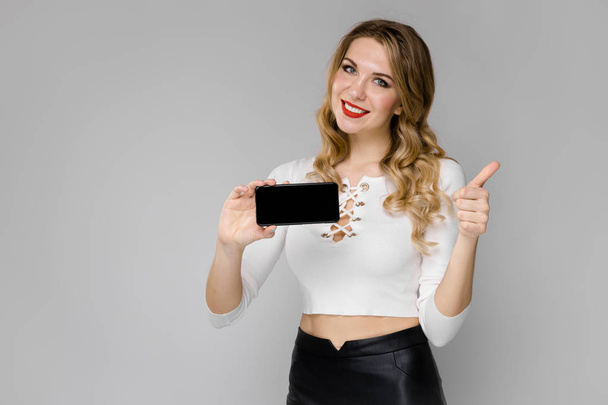 Atractiva joven mujer rubia caucásica en traje casual posando con teléfono inteligente y mostrando diferentes expresiones en la pared blanca en el estudio
.  - Foto, Imagen