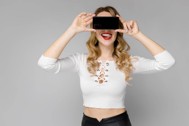 Attractive young caucasian blond woman in casual outfit posing with smartphone and showing different expressions on white wall in studio.  - Zdjęcie, obraz