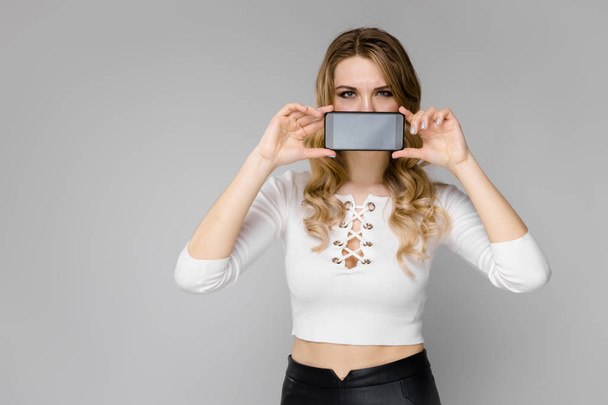 attraktive junge kaukasische blonde Frau in lässigem Outfit posiert mit Smartphone und zeigt verschiedene Gesichtsausdrücke an weißer Wand im Studio.  - Foto, Bild