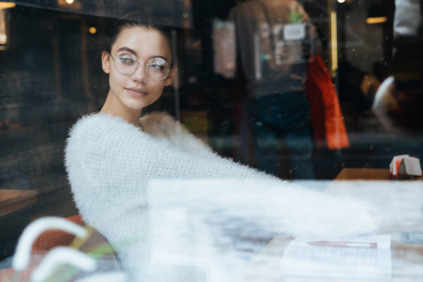 Kallis nuori tyttö freelancer lasit ja valkoinen takki istuu kahvilassa ja katselee ulos ikkunasta
 - Valokuva, kuva