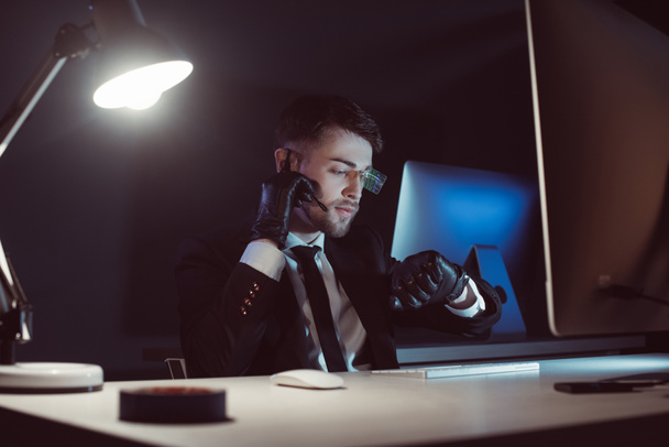 portrét špionážní agent v rukavice s mikrofonem kontrola času u stolu s obrazovkou počítače ve tmě - Fotografie, Obrázek