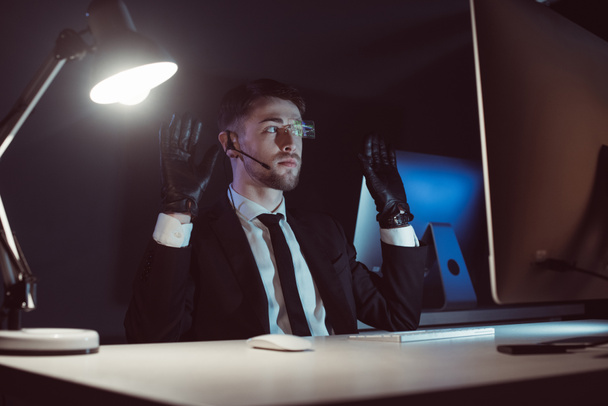 spion agent in handschoenen met handen omhoog zittend aan tafel in donker - Foto, afbeelding