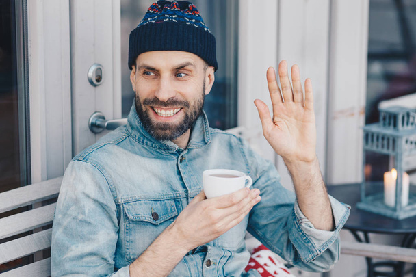 Horizontal portrait of happy male with dark beard and mustache, waves with hand, glad to notice friend, holds cup of tea, wears fashionable clothing. Man greets someone. Hipster guy drinks coffee - Photo, Image