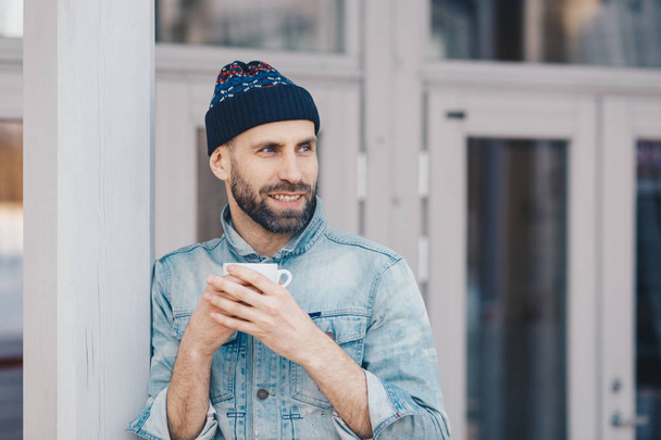 Горизонтальный снимок красивого улыбающегося мужчины счастливо смотрит в сторону, носит шляпу и джинсовую куртку, держит кружку кофе, замечает что-то приятное, отдыхает дома. Человек, образ жизни и отдых
 - Фото, изображение