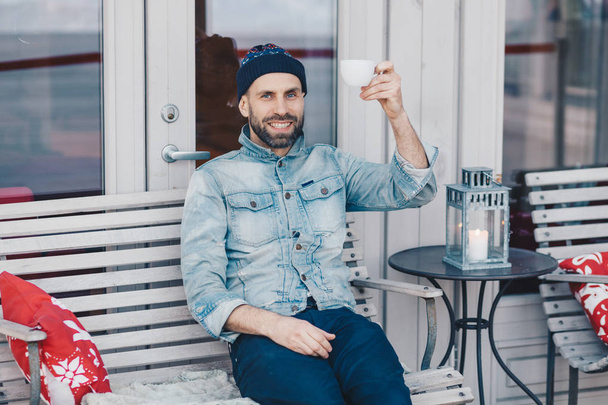 Kuva tyytyväinen sinisilmäinen parrakas mies pukeutunut denim vaatteet, nostaa käsi kuppi kahvia tai teetä, on hyvällä tuulella, lepää ulkona kahvilassa kodikas sisustus ja rauhallinen tunnelma
 - Valokuva, kuva