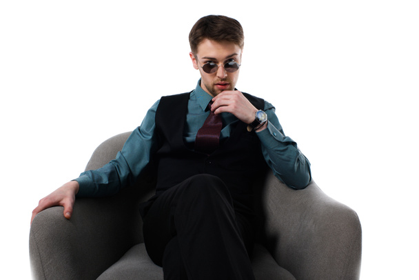portrait of pensive stylish spy in sunglasses sitting in armchair isolated on white - Photo, Image