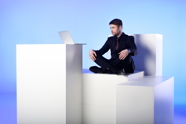 Geschäftsmann mit Laptop sitzt auf weißem Block isoliert auf weißem Block - Foto, Bild