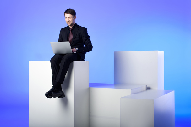 smiling businessman with laptop sitting on white block isolated on white - Photo, Image