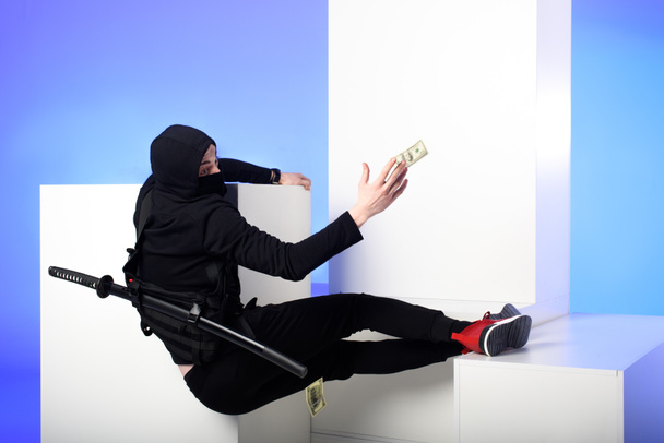 Ninja σε μαύρη ενδυμασία για την αλίευση Δολάριο τραπεζογραμμάτια λευκό μπλοκ που απομονώνονται σε μπλε - Φωτογραφία, εικόνα