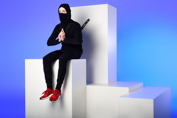 ninja in black clothing with katana behind sitting on white block isolated on blue - 写真・画像