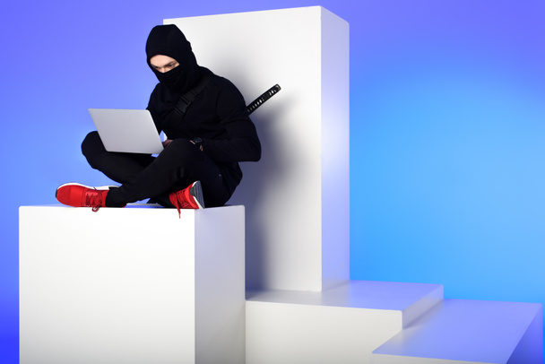 ninja in black clothing using laptop while sitting on white block isolated on blue - Фото, зображення