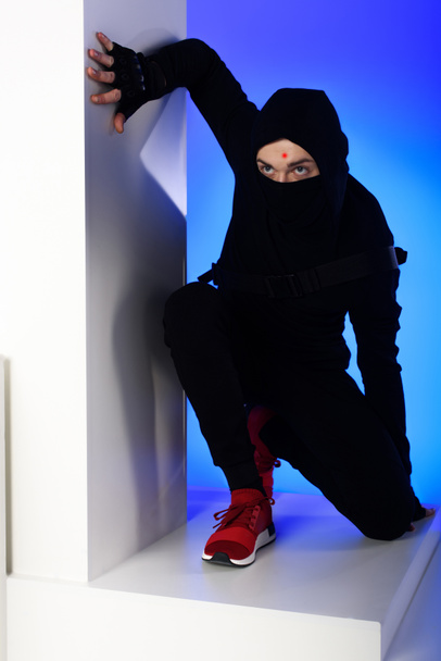 ninja in black clothing with katana behind standing on white block isolated on blue - Photo, Image