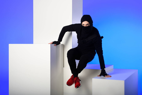 ninja in black clothing with katana behind getting across on white block isolated on blue - Photo, Image