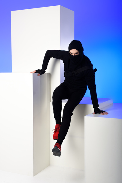 Ninja in schwarzer Kleidung mit Katana dahinter auf weißem Block isoliert auf blauem - Foto, Bild