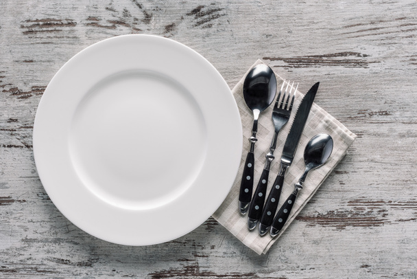 valkoinen levy ja ruokailuvälineet lautasliinalla puupöydällä
 - Valokuva, kuva