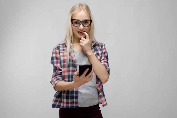 Szép kaukázusi szőke tizenéves lány kockás ing és a szemüveg, mutatja a különböző érzelmi kifejezések stúdióban szürke falon telefon használata közben. - Fotó, kép