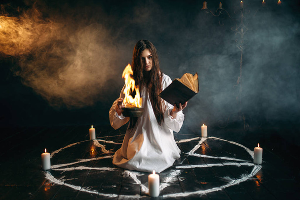 young woman in white shirt sitting in the center of pentagram circle with candles and reading a spell, dark magic ritual, occultism - Photo, Image