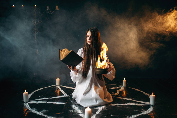 Μάγισσα σε λευκό πουκάμισο ανάγνωση ξόρκι, πεντάγραμμο κύκλο με κεριά, σκοτεινό μαγικό τελετουργικό. Αποκρυφισμός και εξορκισμός - Φωτογραφία, εικόνα