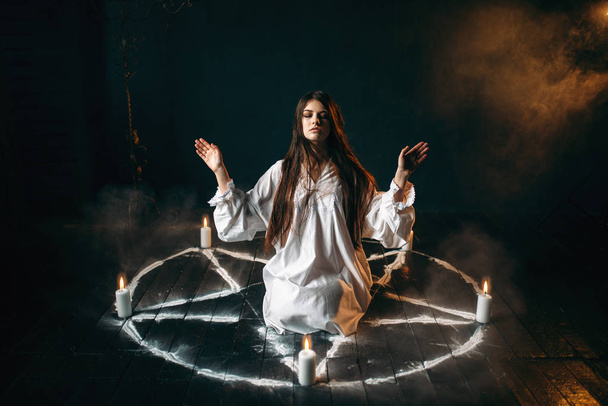 молодая женщина в белой рубашке, сидящая в центре пентаграммного круга со свечами, ритуал черной магии, черный деревянный пол
 - Фото, изображение