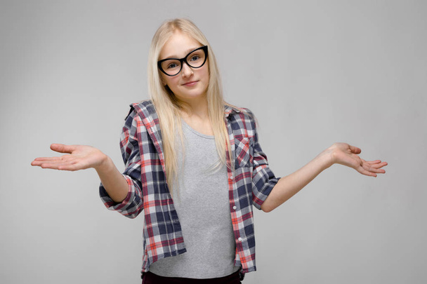 Hübsches kaukasisches blondes Teenie-Mädchen in kariertem Hemd und Brille mit unterschiedlichen Gefühlsausdrücken an grauer Wand im Studio. Hände verteilen. - Foto, Bild
