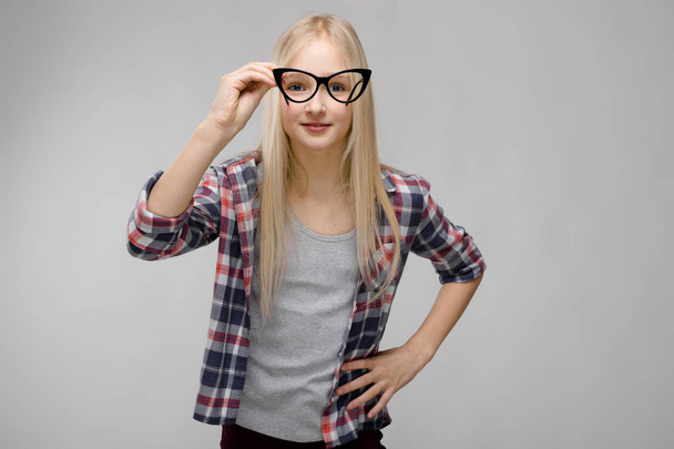 Menina adolescente loira muito caucasiana em camisa quadriculada mostrando diferentes expressões emocionais na parede cinza no estúdio. Olhando para a câmera através de óculos
. - Foto, Imagem