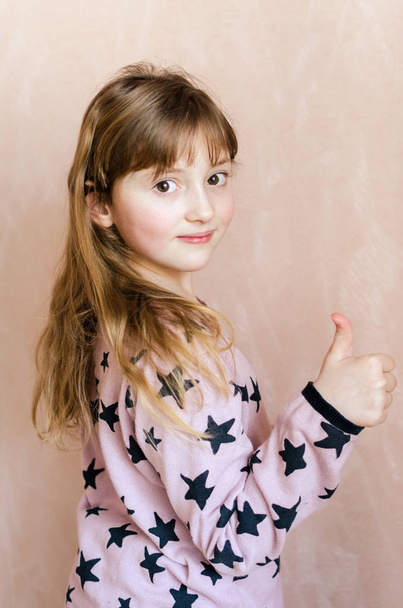 Little girl with long dark blonde hair and a cute smile on face makes thumbs-up gesture. Stylish clothes. Pink background. - Photo, Image