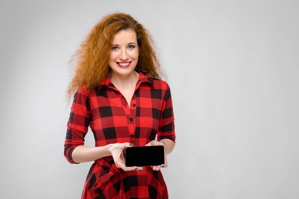 Όμορφη κοκκινομάλλα σγουρά καυκάσιος γυναίκα σε κόκκινο καρώ φόρεμα δείχνουν διαφορετικές συναισθηματικές εκφράσεις στον λευκό τοίχο στο studio. Χαμογελώντας και δείχνοντας κενό smartphone. - Φωτογραφία, εικόνα
